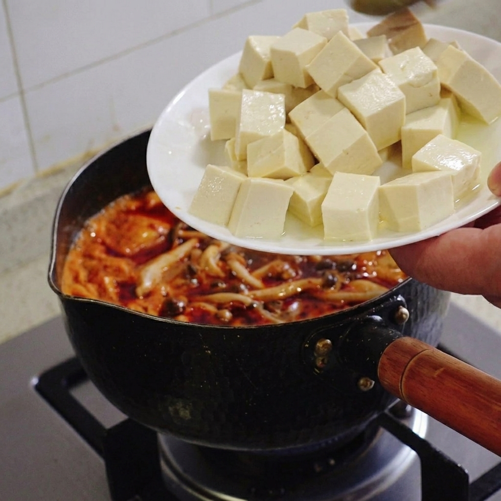 A hand holding a white plate and sliding neat cubes of soft white tofu into a simmering pot of red kimchi stew.