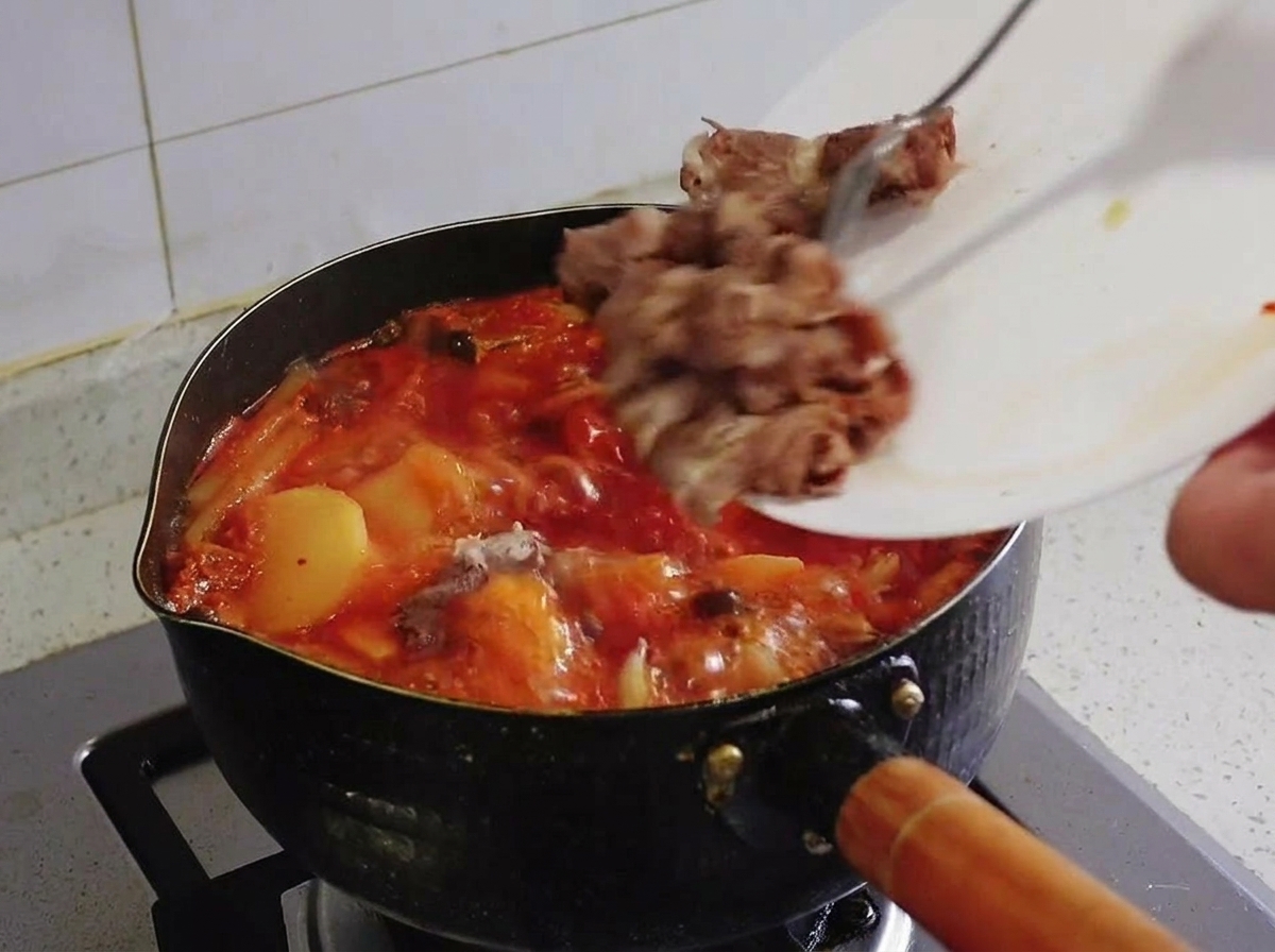 Blanched fatty beef pieces being slid from a white plate into a boiling pot of red kimchi stew.