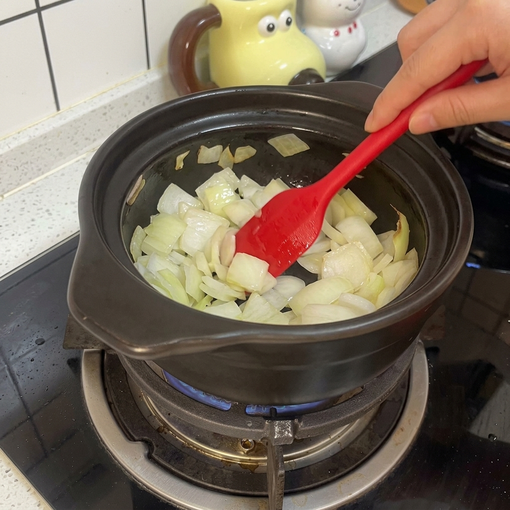 A red silicone spatula stirring chopped white onions in a black clay pot over a gas stove flame.