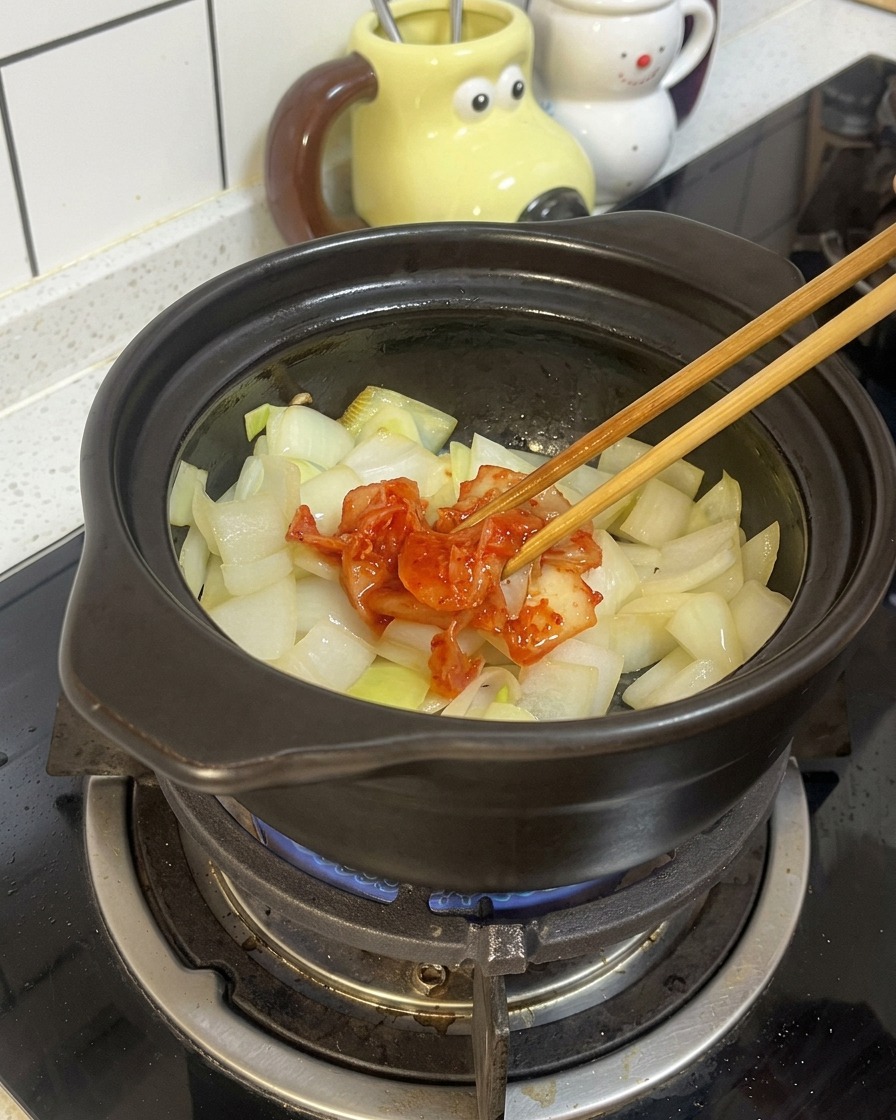 Wooden chopsticks placing a portion of red, fermented kimchi on top of sautéed onions in a black clay pot.
