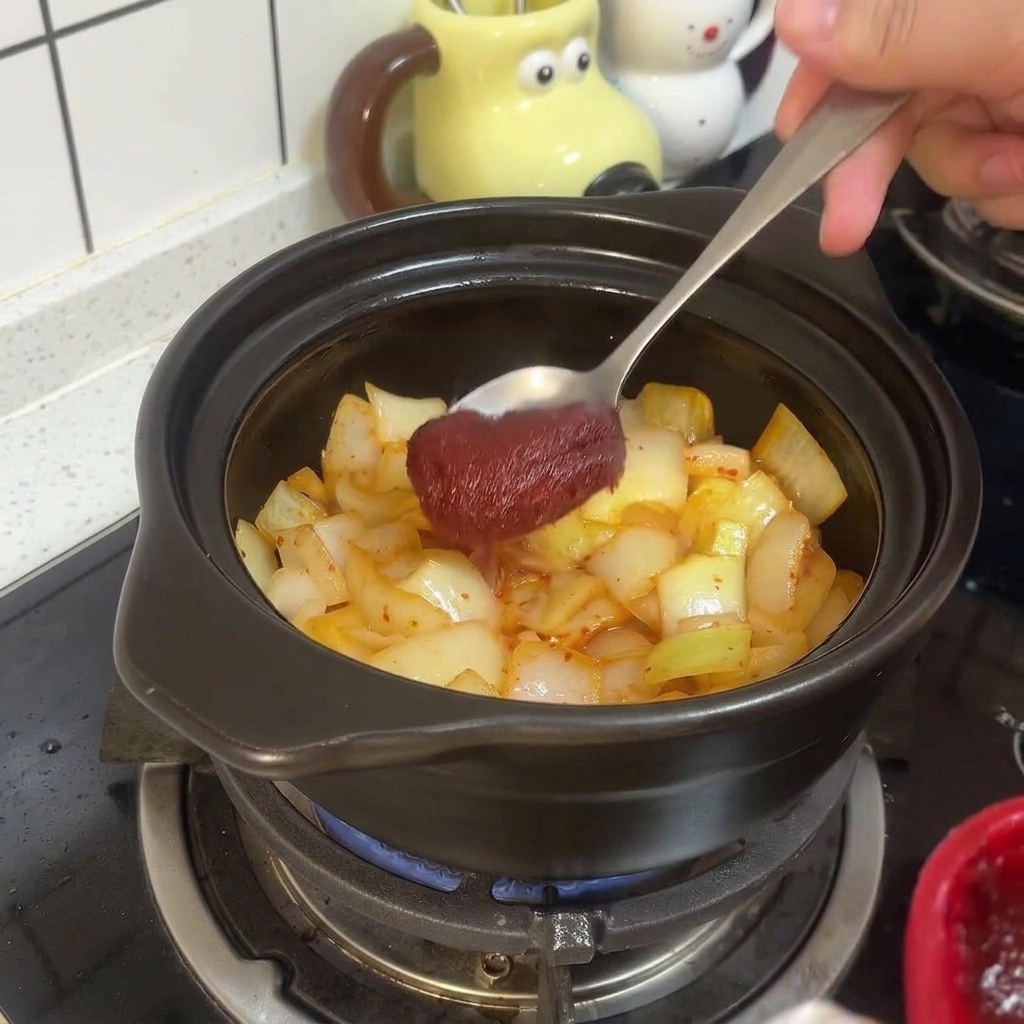 A metal spoon mixing thick red Korean chili paste into the sautéed kimchi and onions inside the clay pot.