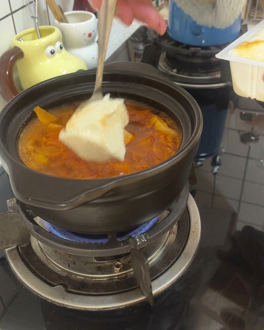 A spoon scooping large pieces of soft tofu into a bubbling red broth in a clay pot.