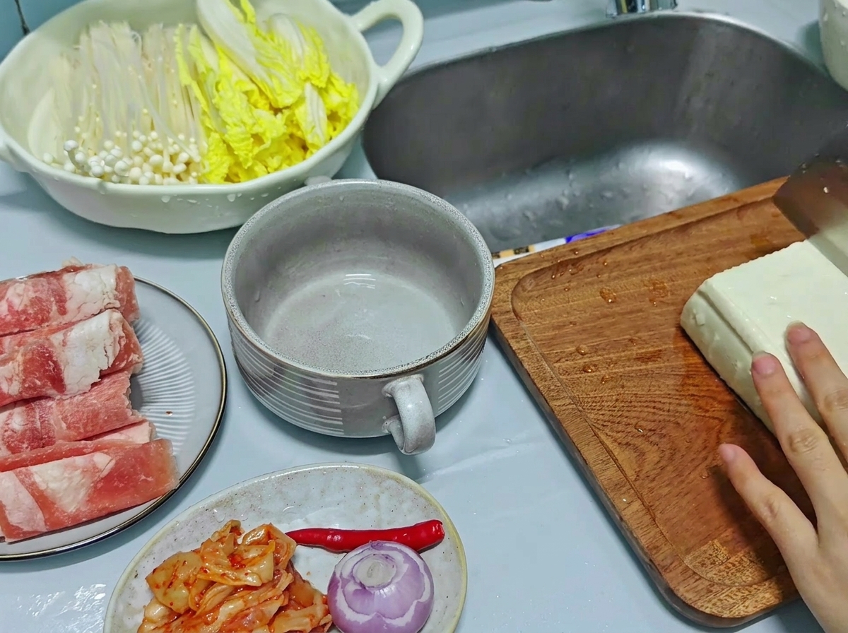 A close-up view of a hand using a kitchen knife to slice a large block of white tofu into thick rectangular pieces on a wooden cutting board.