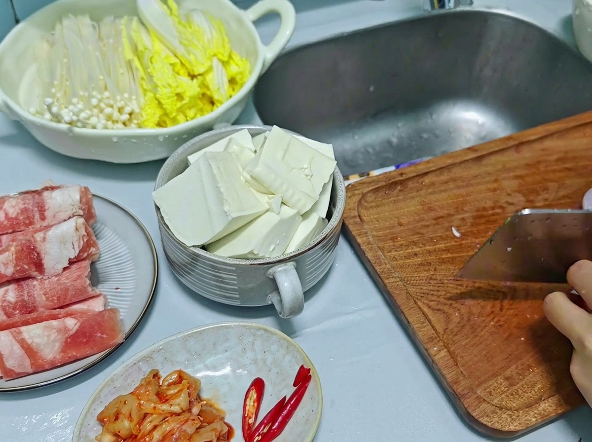 A kitchen cleaver being used to chop red onions and red chili peppers on a wooden cutting board, with sliced tofu visible in a side bowl.
