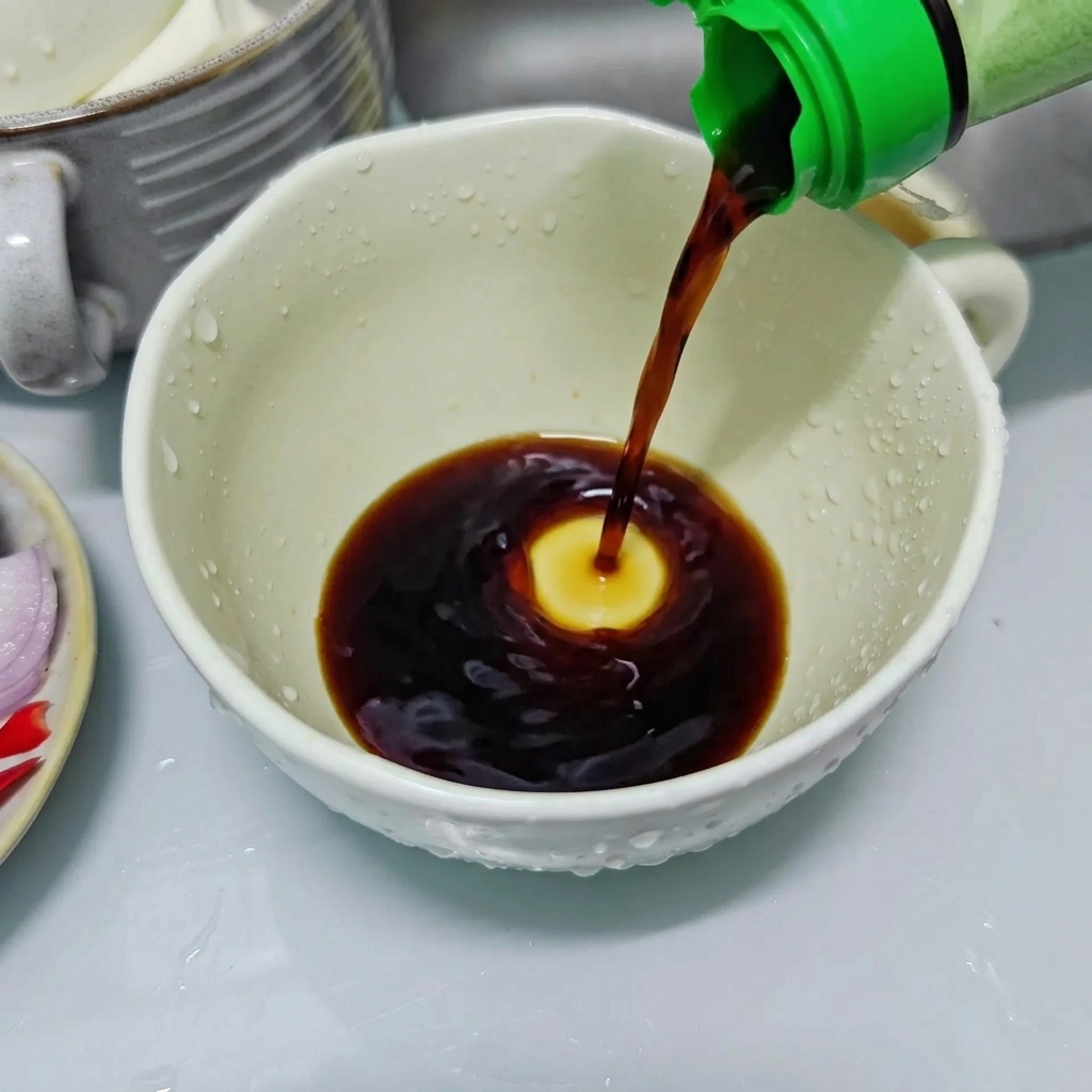 Dark soy sauce being poured from a green-capped bottle into a white ceramic cup to prepare the seasoning sauce.