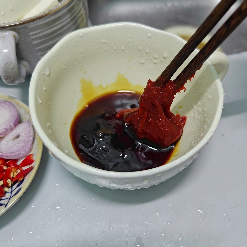 Wooden chopsticks holding a large dollop of thick red Korean chili paste over a white ceramic bowl filled with dark savory sauces.