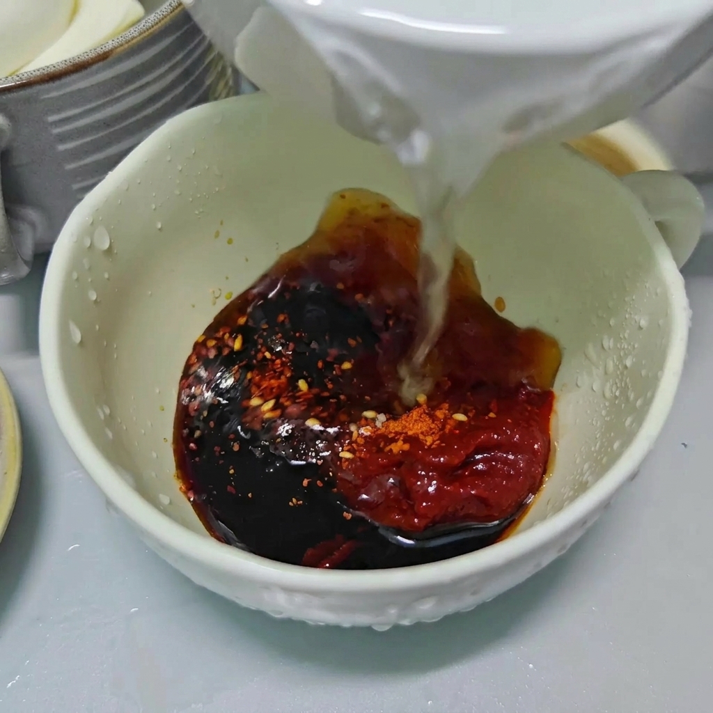 Clear water being poured into a white bowl filled with dark soy sauce, red chili paste, and chili flakes.