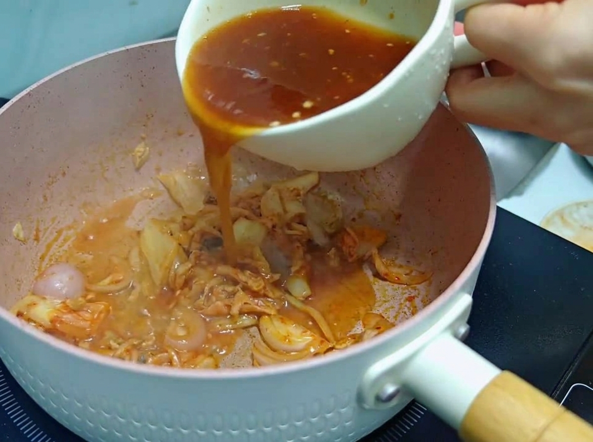 A person pouring a reddish-brown mixed sauce from a white mug into a pink cooking pan containing sautéed kimchi and onions.