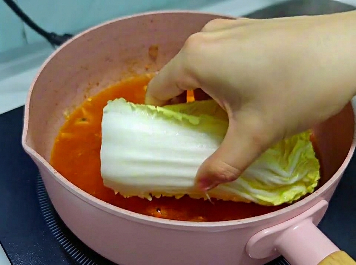 A hand carefully placing a large piece of fresh napa cabbage into a pink pan filled with bubbling kimchi soup broth.