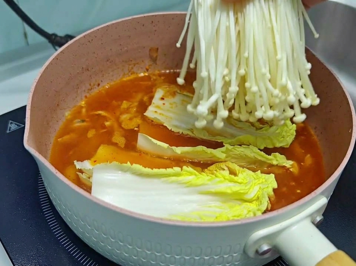 A close-up of a hand lowering a fresh, clean bundle of white enoki mushrooms into a pink cooking pot containing a rich orange-red kimchi soup base and napa cabbage.