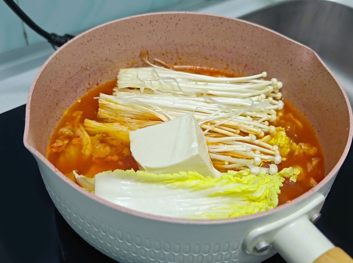 A large block of fresh white tofu resting on top of enoki mushrooms and cabbage inside a pot of orange kimchi soup.