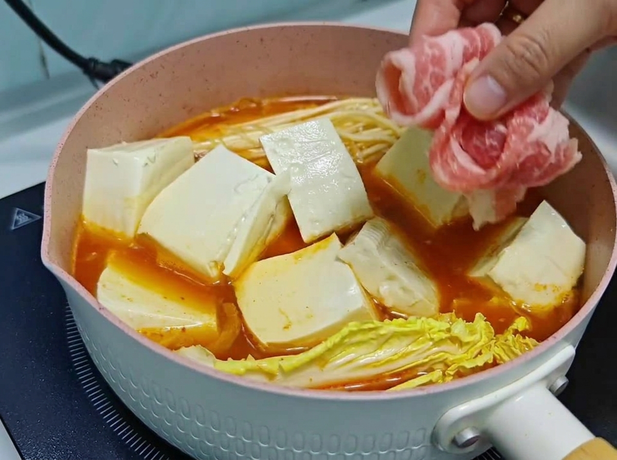 A hand carefully layering thin rolls of raw beef over tofu cubes and vegetables in a simmering kimchi soup pot.