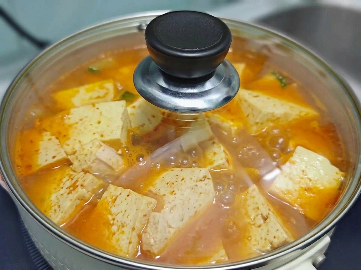 A clear glass lid covering the pot of bubbling Korean kimchi beef tofu soup as it simmers on the stove.