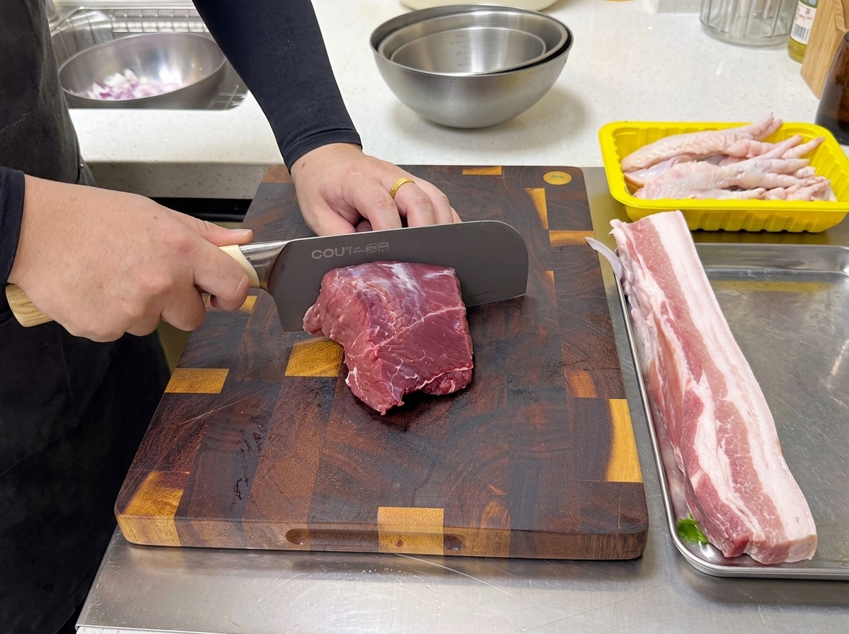 A chef using a large professional knife to slice a piece of raw red beef chuck eye roll on a wooden cutting board.