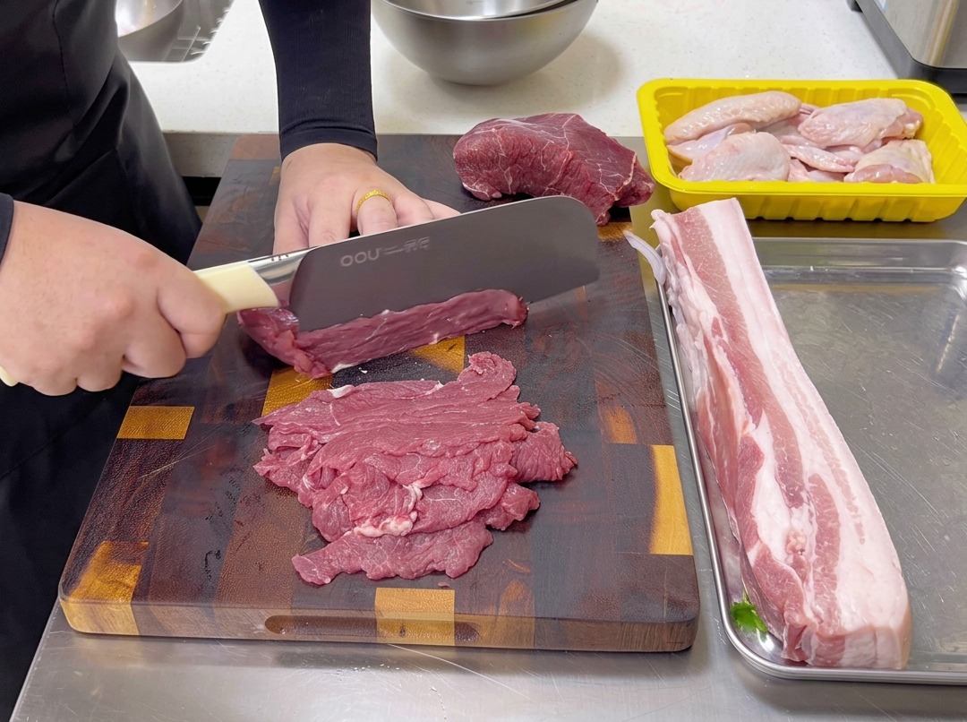 Thinly slicing raw beef against the grain on a wooden cutting board with a chefs knife.