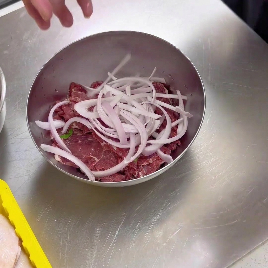 Slices of fresh raw beef inside a metal mixing bowl topped with a handful of freshly sliced white onions.