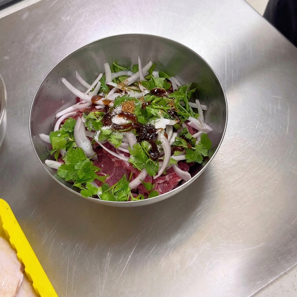 A metal bowl containing raw sliced beef, onions, and cilantro topped with dry spices including cumin powder, cumin seeds, salt, and sugar.