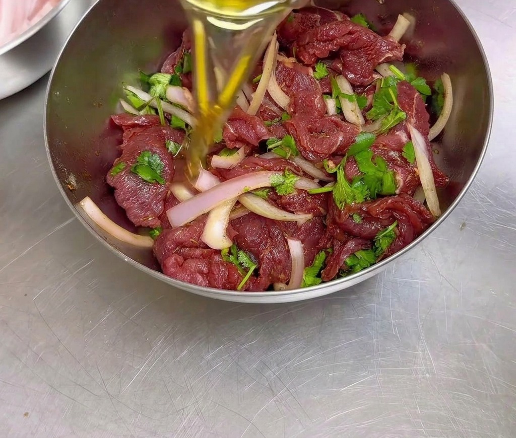 A person pouring cooking oil into a stainless steel bowl filled with seasoned marinating beef, sliced onions, and cilantro.