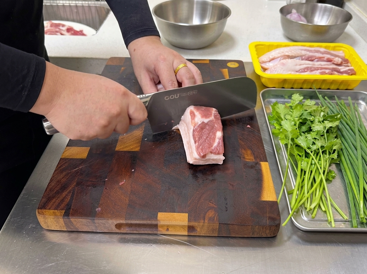 A person using a large kitchen knife to slice a block of raw, layered pork belly into thin strips on a cutting board.