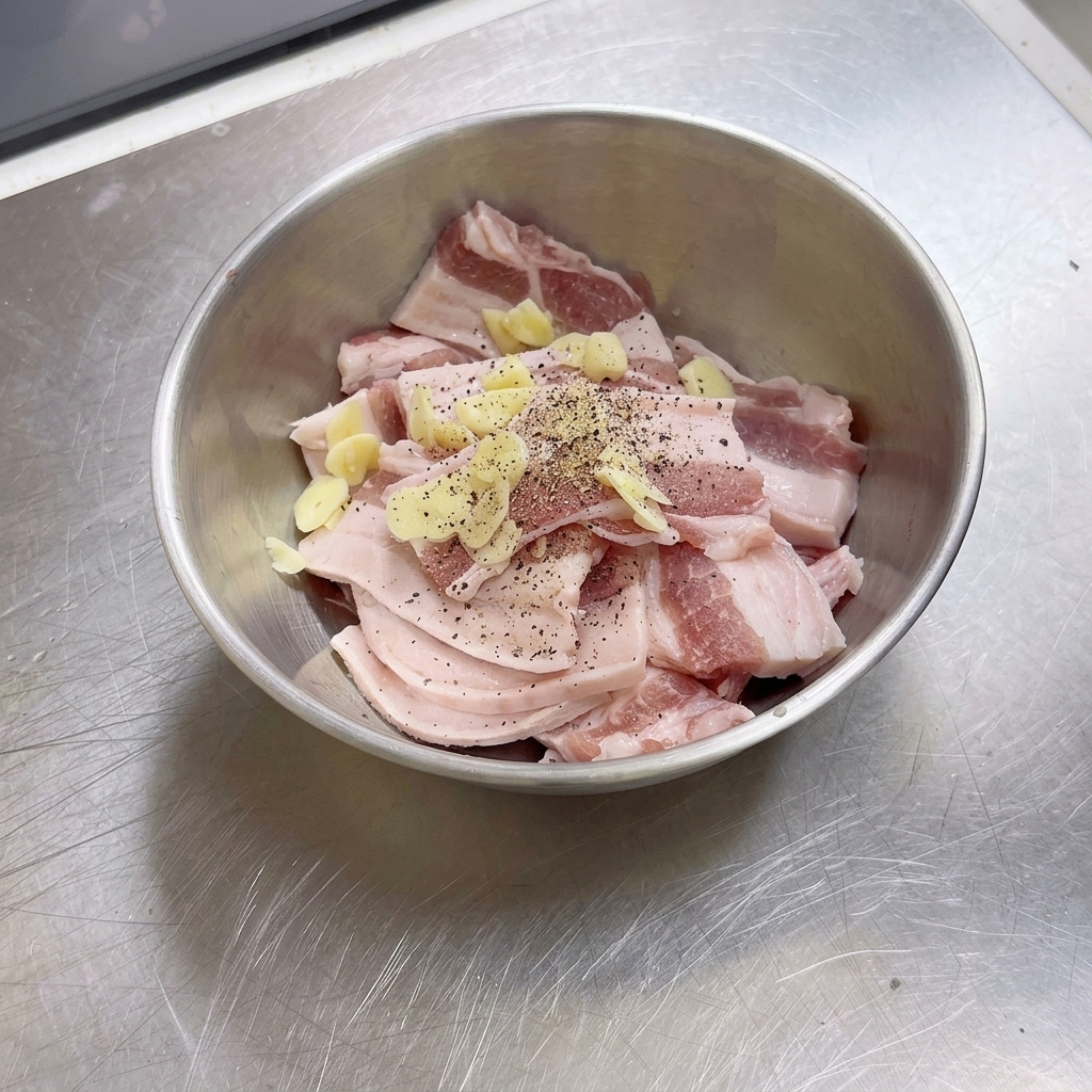 A stainless steel bowl filled with raw pork belly slices, garlic pieces, and freshly cracked black pepper.