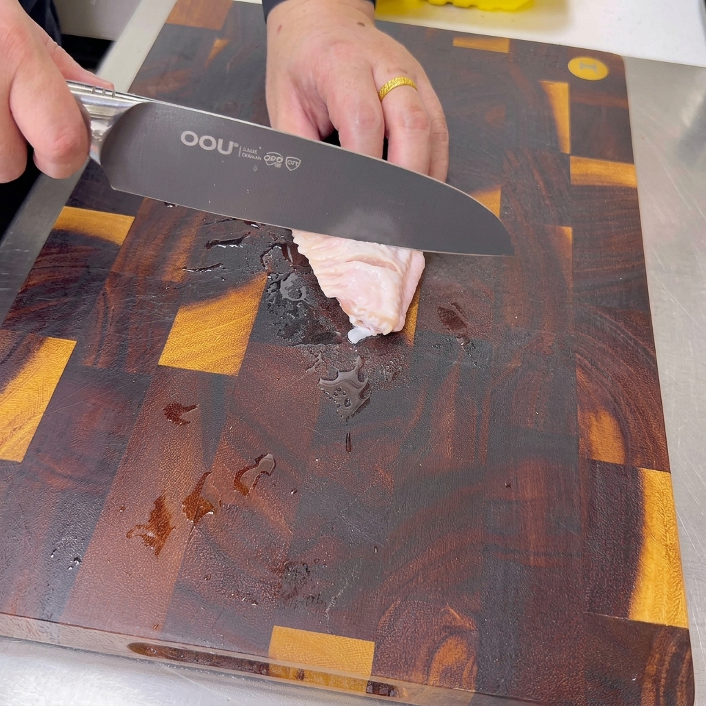 A sharp knife cutting small diagonal slits into a raw chicken wing on a dark wooden cutting board.