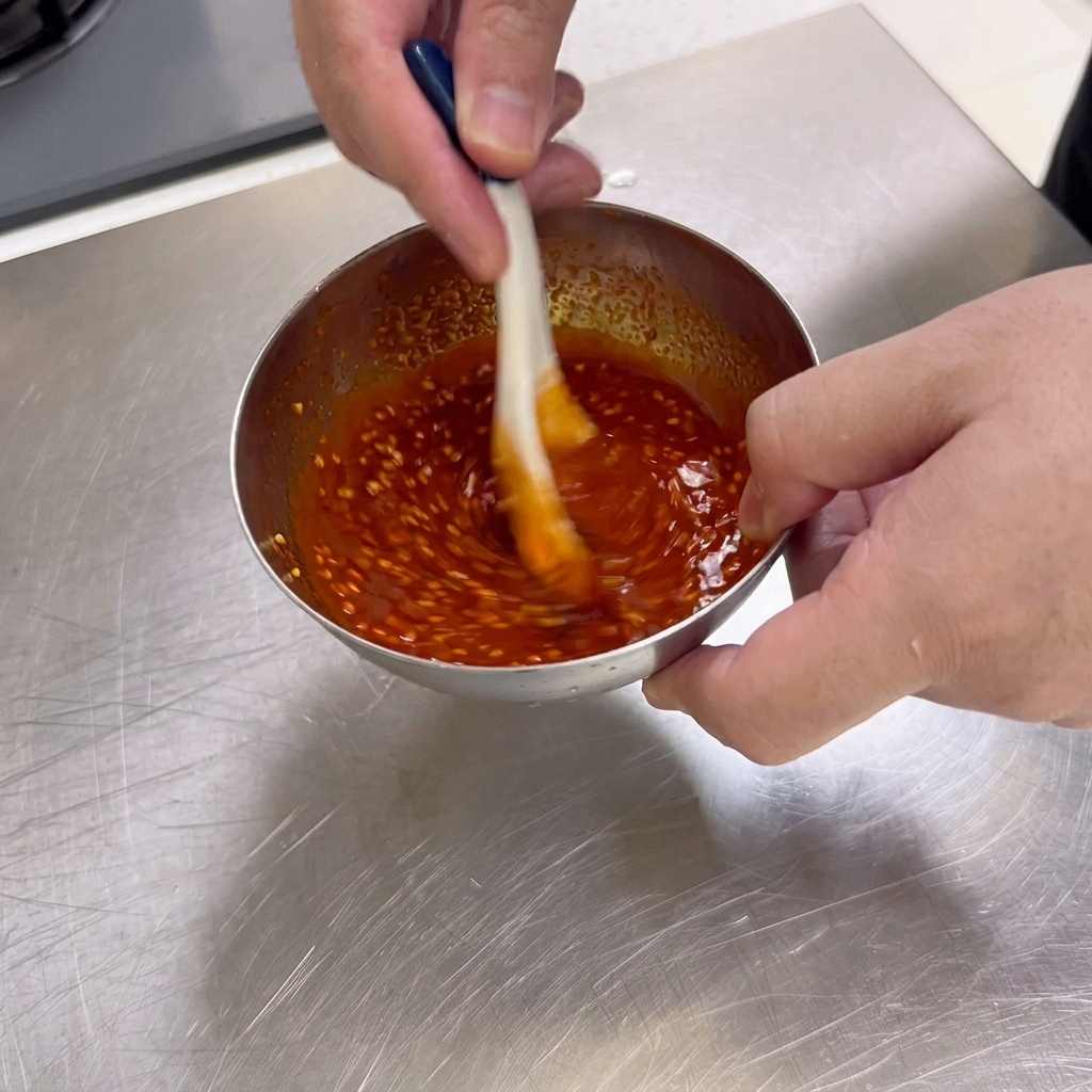 A thick, deep red barbecue dipping sauce filled with sesame seeds being stirred with a spoon in a small stainless steel bowl.