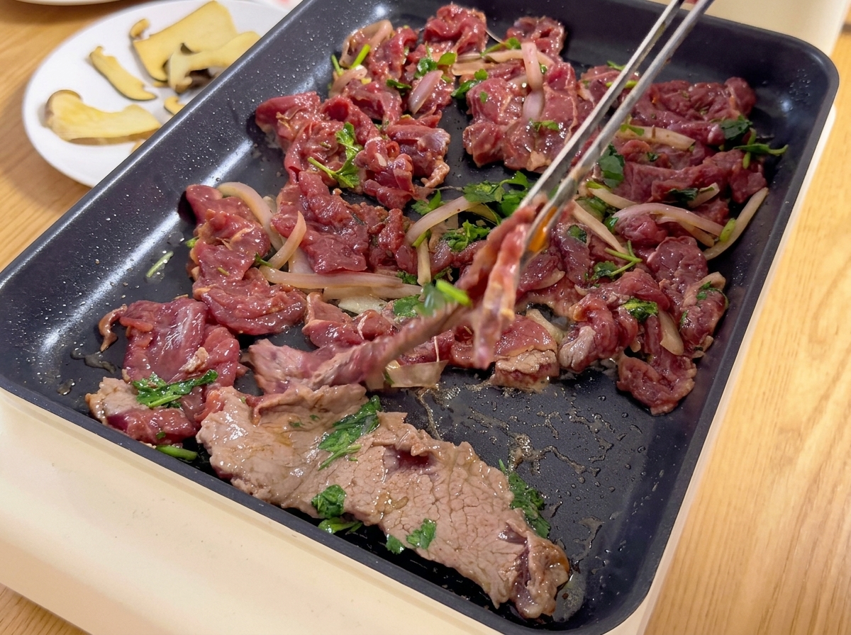 Thinly sliced marinated beef mixed with onions and cilantro being placed onto a hot electric griddle with tongs.