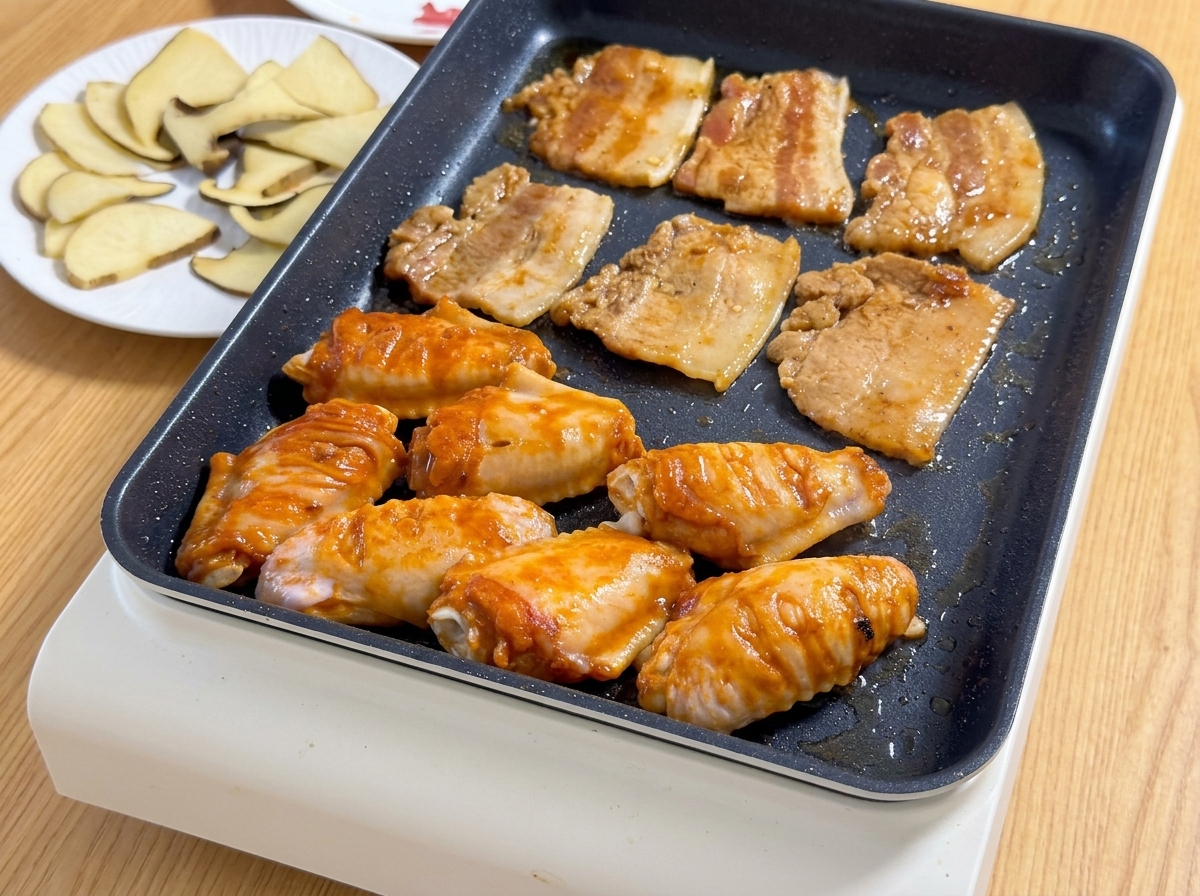 Marinated pork belly slices and orange Orleans chicken wings sizzling together on a hot electric griddle.