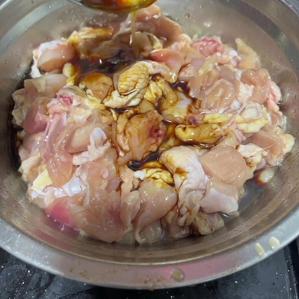 Cooking wine being poured from a stainless steel spoon into a bowl containing raw chicken chunks and dark sauces.