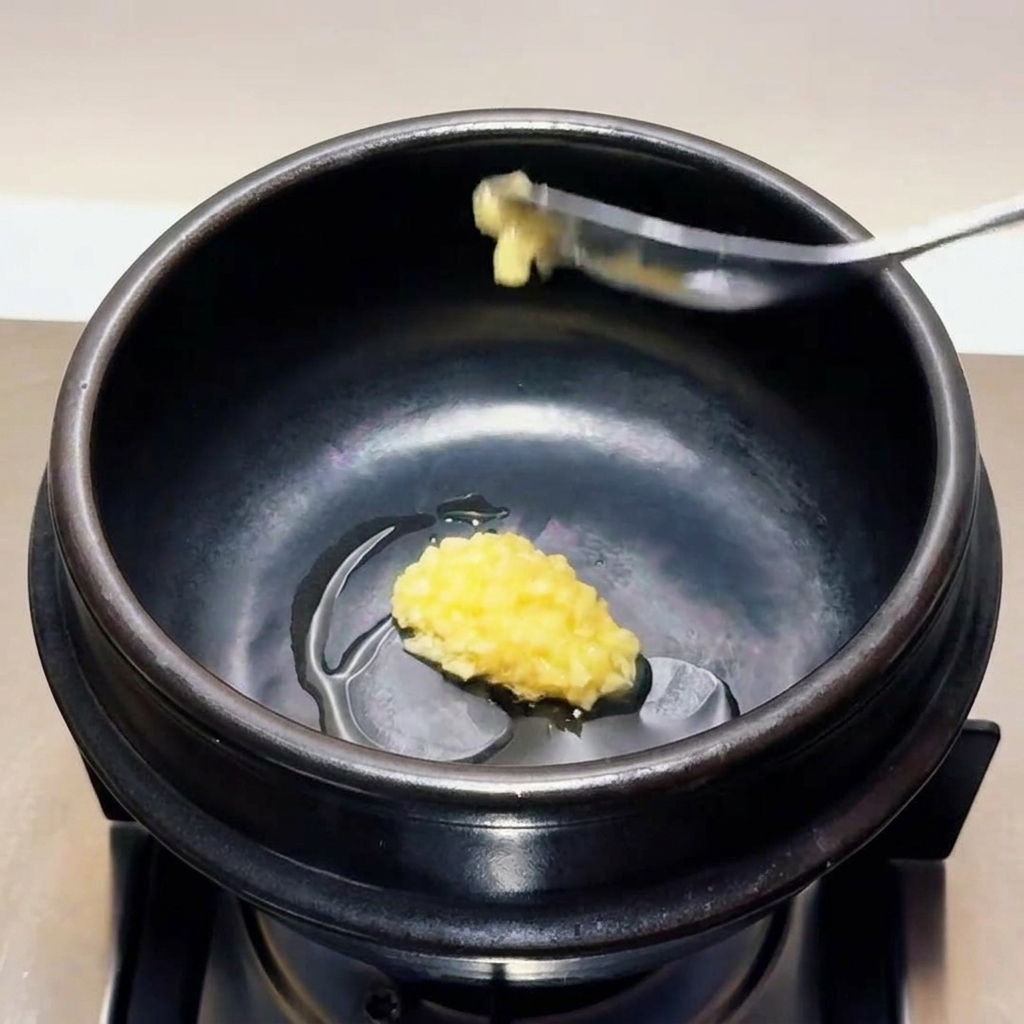 A spoonful of finely minced garlic being lowered and placed into the heated oil inside a black stone cooking pot.