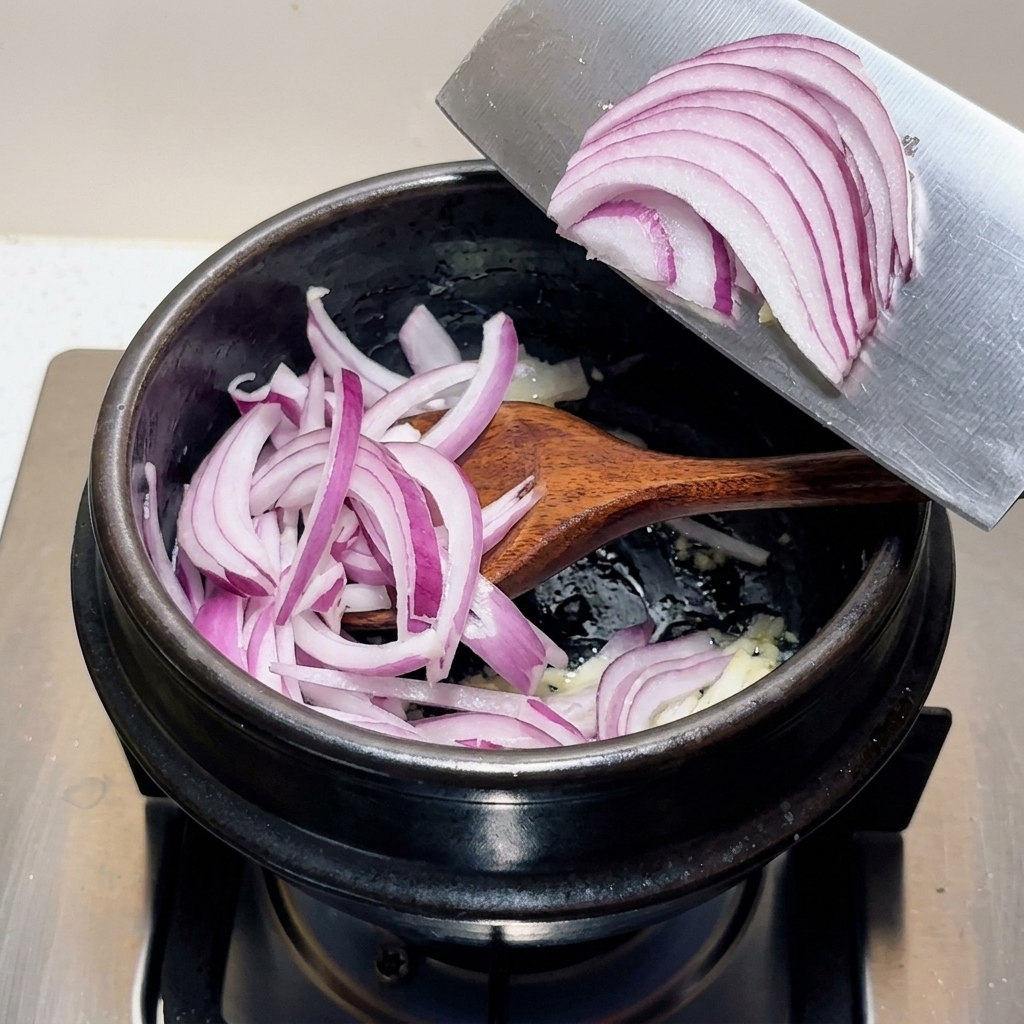 A heap of sliced purple red onions being transferred from a wide metal blade into a black cooking pot containing aromatic ingredients.
