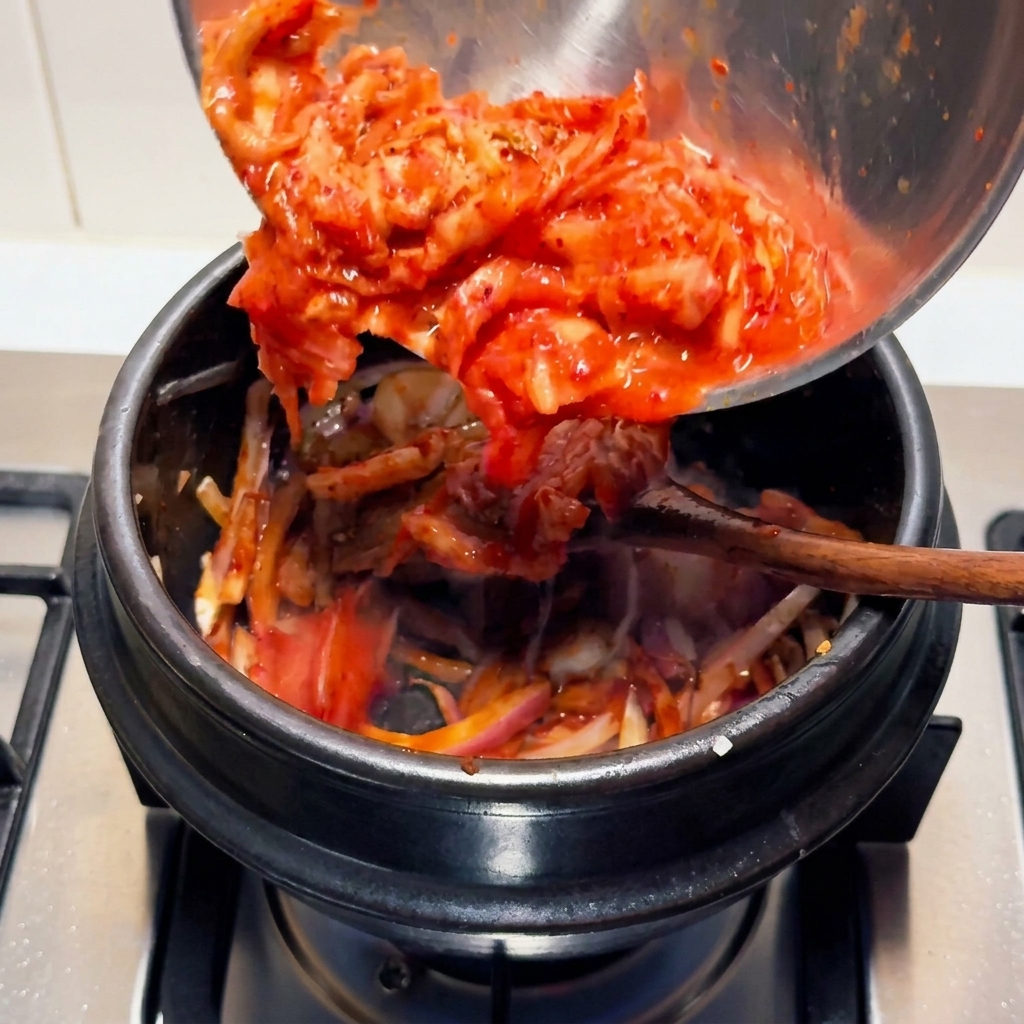 Bright red chopped sour kimchi being poured from a stainless steel bowl into a black stone cooking pot on a stovetop.