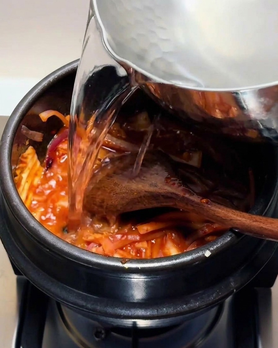 Clear hot water being poured from a metal pot into a black stone pot containing the red kimchi mixture.