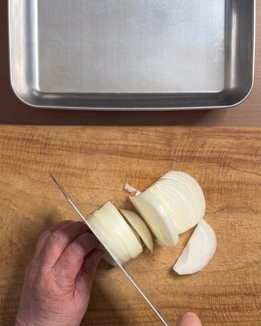 Hands using a knife to slice a white onion into thick strips on a wooden cutting board, with a metal tray nearby.