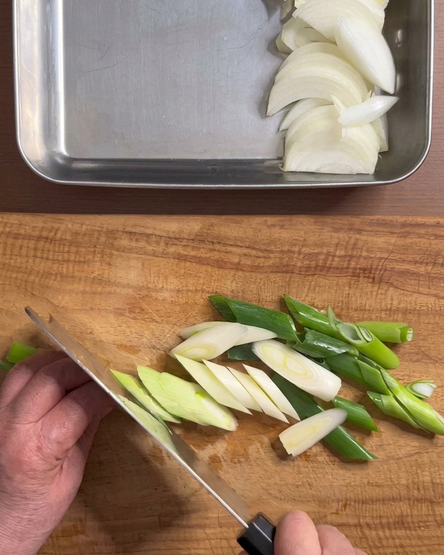 Hands holding a knife slicing a fresh green onion diagonally on a wooden cutting board.