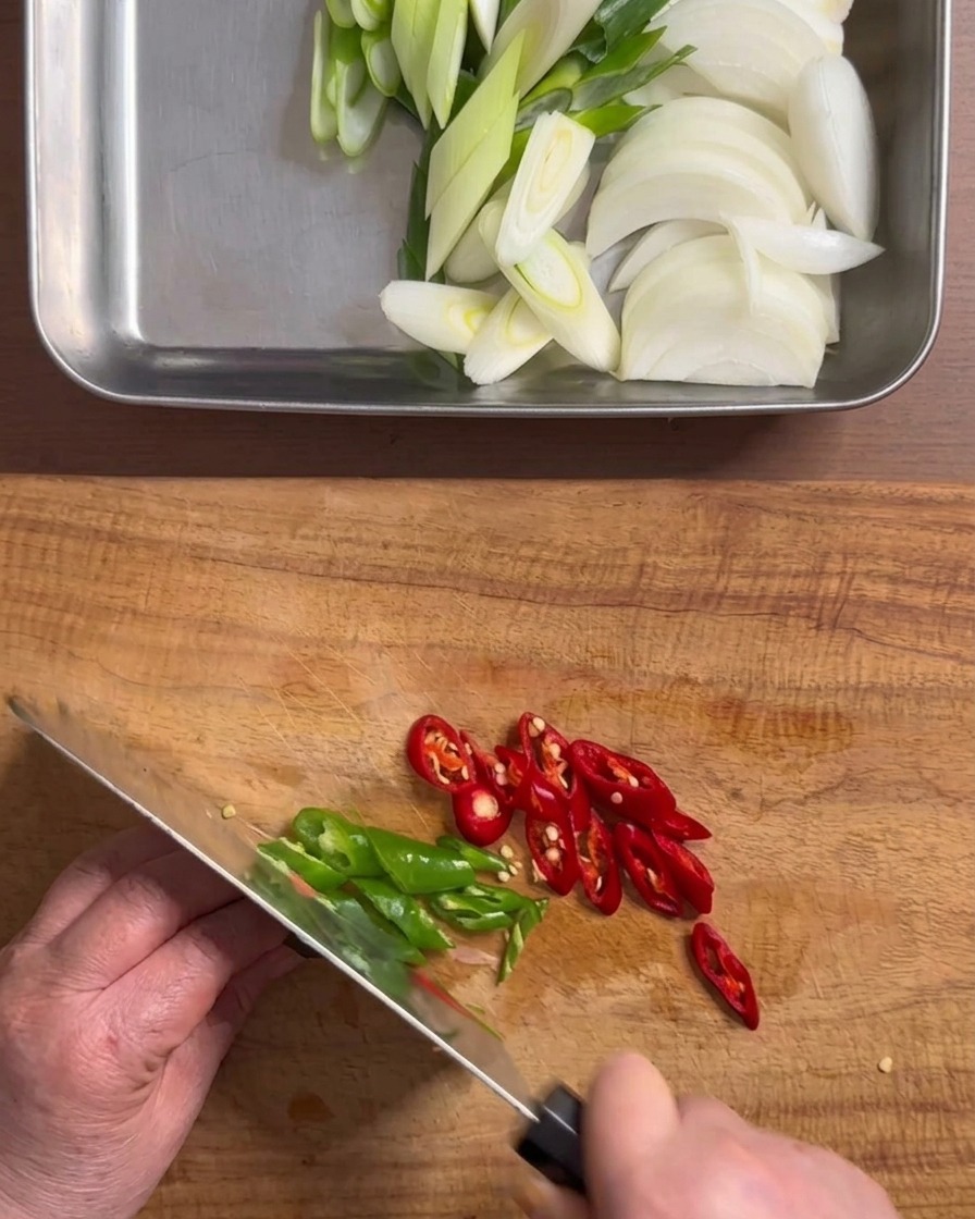 Hands using a knife to chop green and red chili peppers into rings on a wooden cutting board.