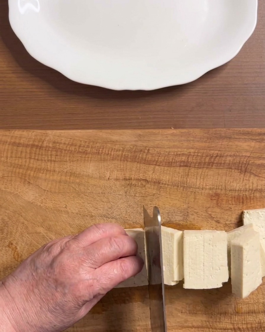 Hands slicing a block of firm white tofu into even rectangular pieces on a wooden cutting board.