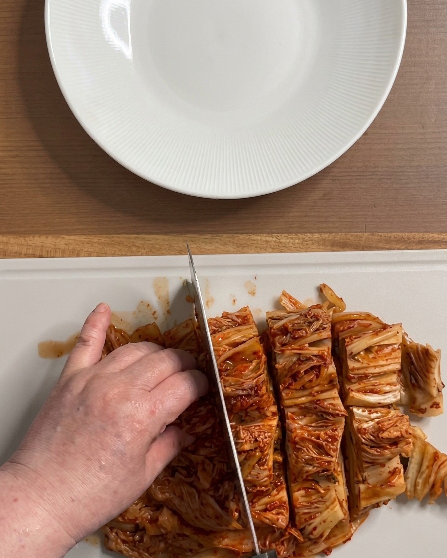 Hands cutting a large, vibrant red block of fermented kimchi into bite-sized sections on a white cutting board.