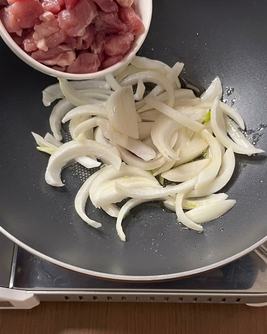 Raw pork pieces being added from a small bowl to a pan with sliced onions and oil.