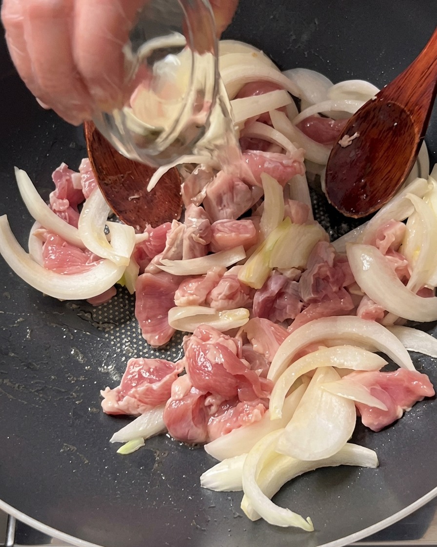 Cooking wine being poured from a small glass bowl onto pork and onions in a pan.