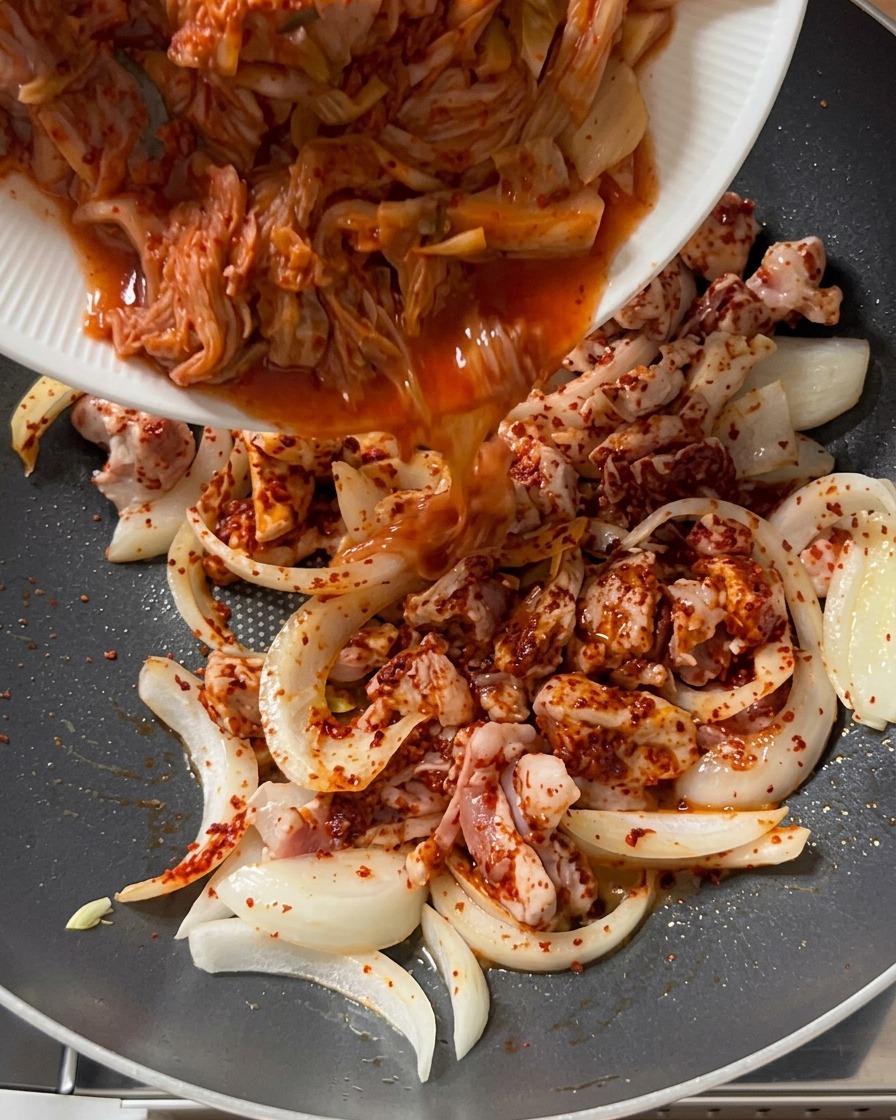 Chopped kimchi being poured from a white plate into a pan containing seasoned pork and onions.