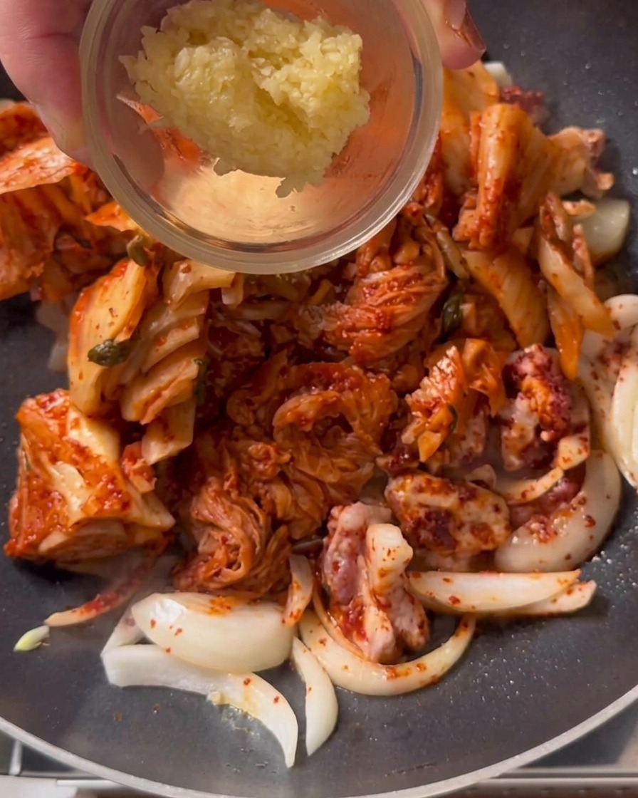 A spoonful of minced garlic being added to a pan of stir-frying kimchi and pork.