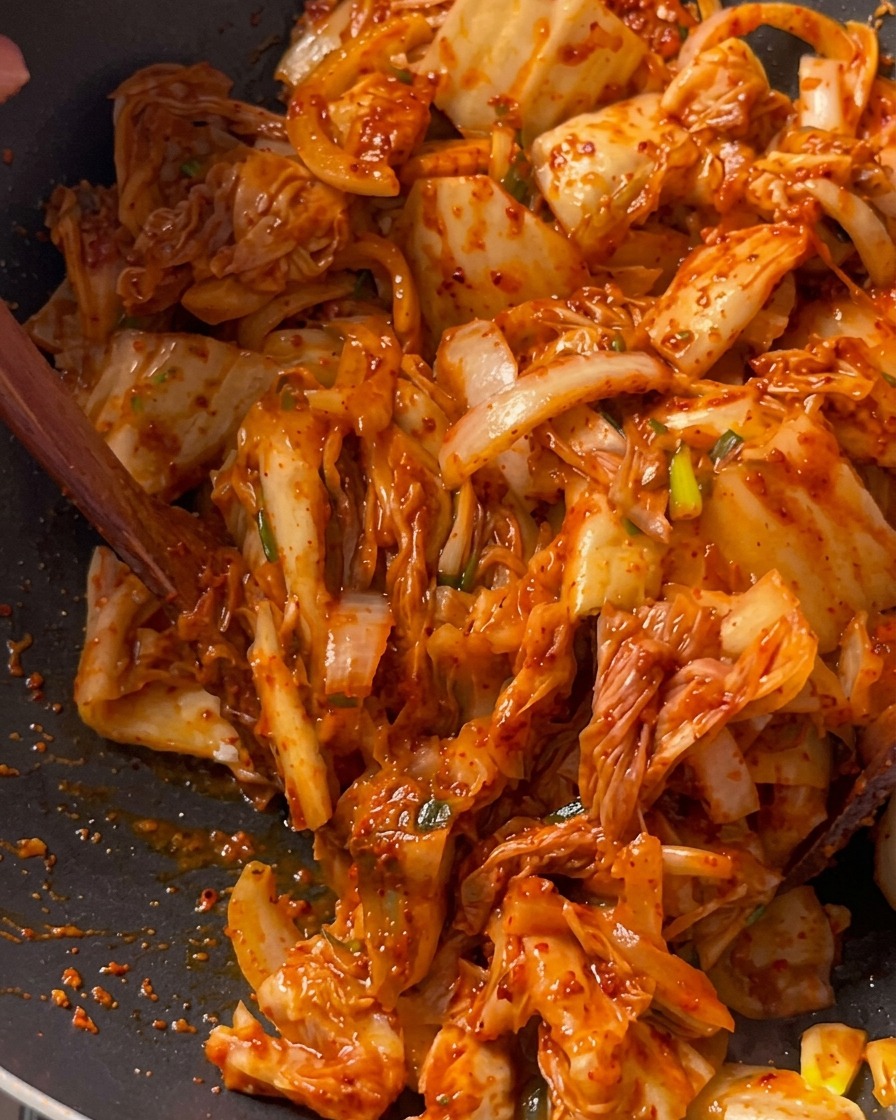 Kimchi pieces being stir-fried together with pork and onions in a deep black wok using a wooden spatula