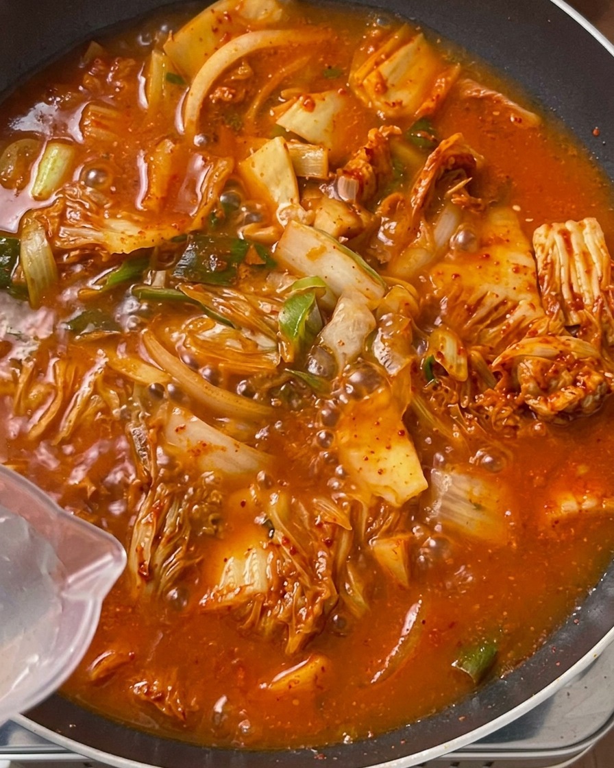 Clear water being poured from a transparent plastic pitcher into a large pan filled with a bubbling, bright orange-red stir-fried kimchi and meat mixture.