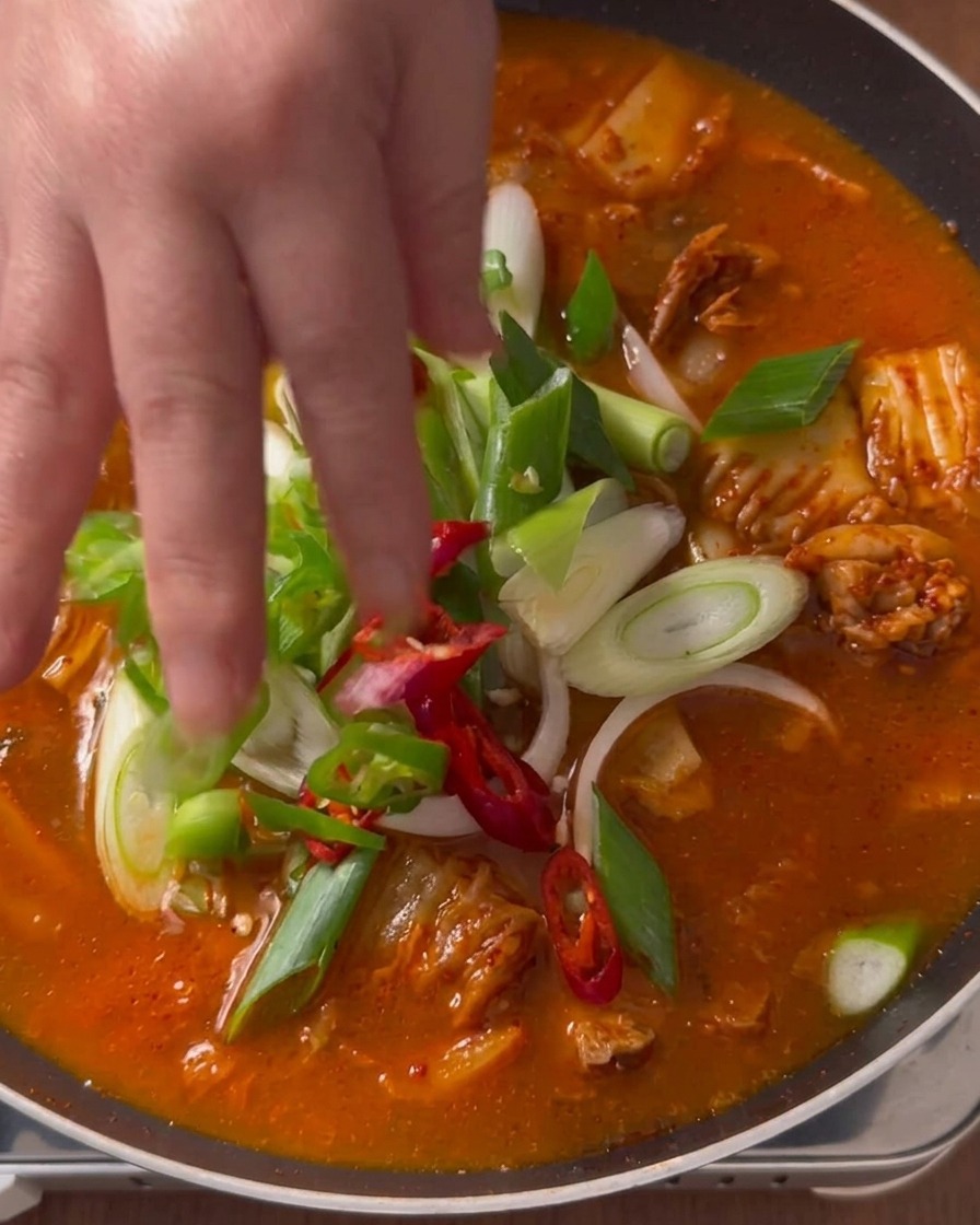 A hand placing freshly cut green scallions, green chilies, and red pepper slices on top of a simmering red kimchi stew in a pan.