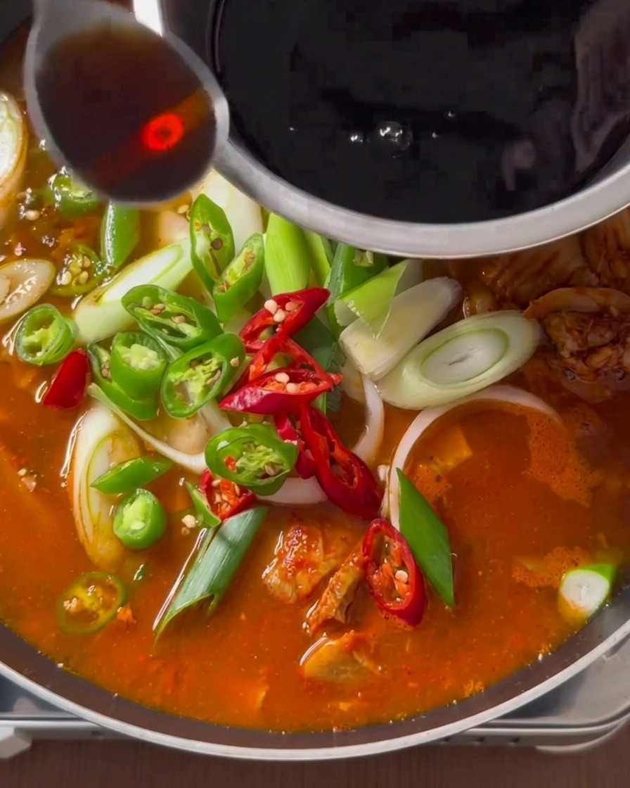 A spoonful of dark liquid seasoning being poured from a stainless steel bowl over a simmering kimchi stew topped with fresh peppers and scallions.