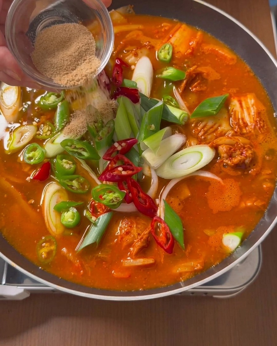 Dry brown beef seasoning powder being sprinkled from a small clear glass dish onto the surface of a simmering red stew filled with peppers and scallions.