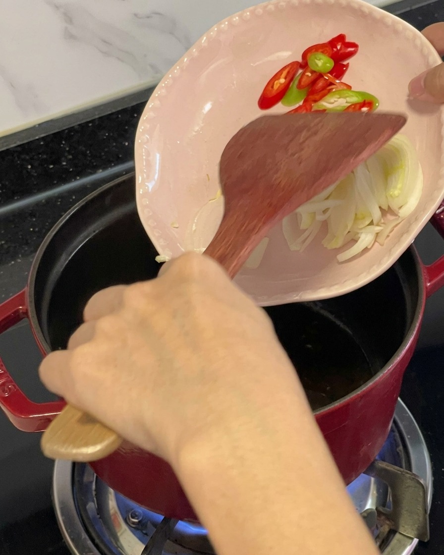 Sliced red and green chilies and aromatics being scraped from a small pink plate into an oiled red pot using a wooden spatula.