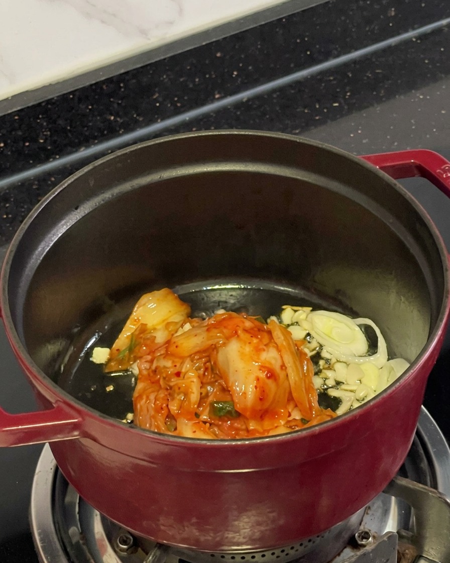 Chopped red kimchi, sliced garlic, and scallions cooking in oil inside a pot.