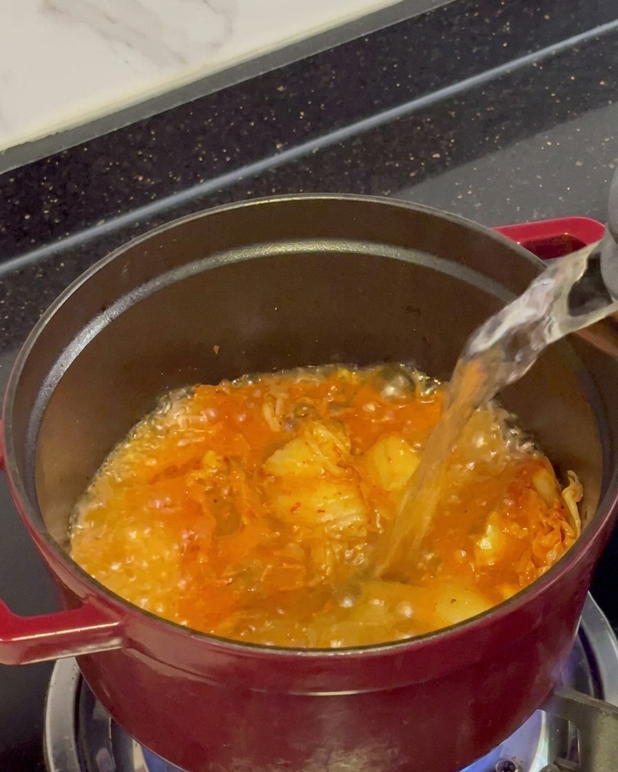 Clear boiling water being poured from a kettle into a red pot filled with bubbling, sautéed kimchi.